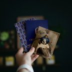 A hand holds three decorative cards or art prints against a dark, blurred background. The set includes a blue patterned card, a dark blue card with gold lettering, and a tan card featuring an illustrated figure with a deer head and Arabic calligraphy.