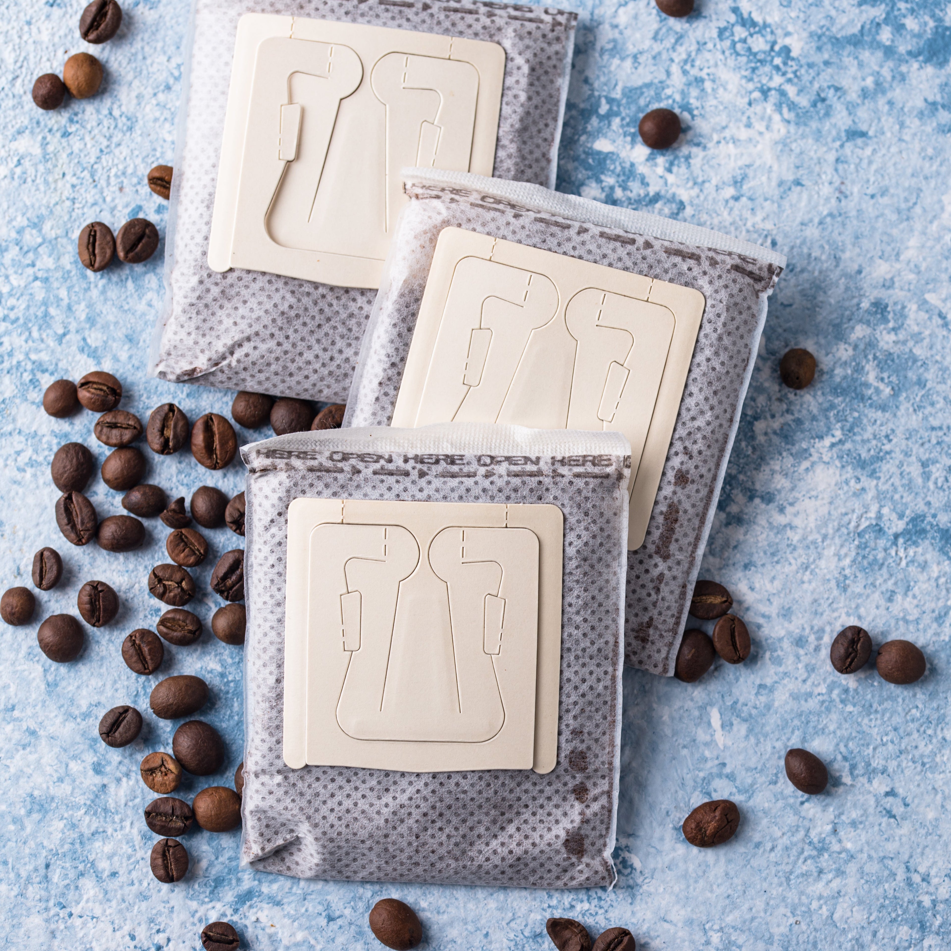 Three coffee filter packages on a blue textured surface with scattered coffee beans.