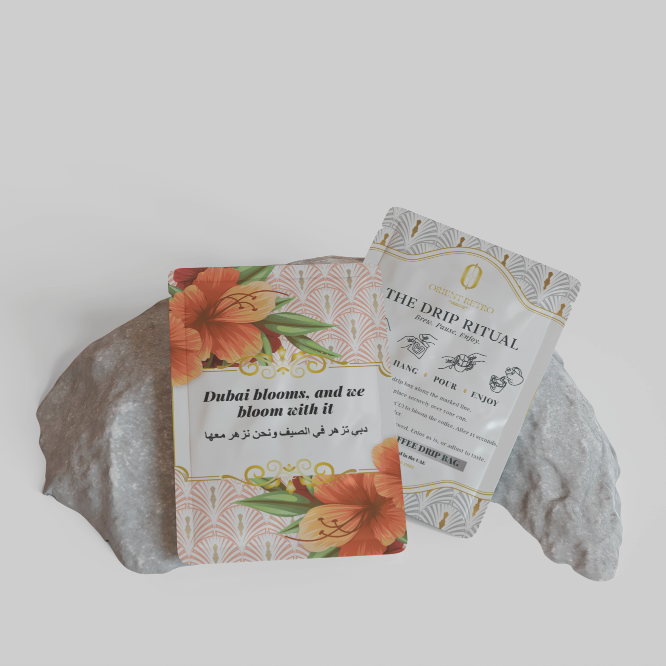 Two small printed cards or sachets displayed on a gray stone against a light gray background. The front card has orange floral artwork and the text 'Dubai blooms, and we bloom with it.