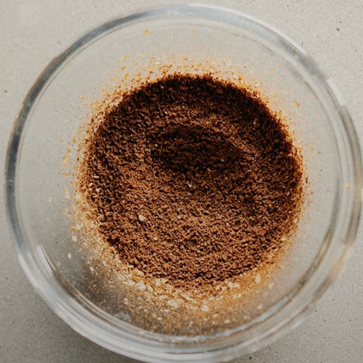 A top-down view of a brown spice or powder mixture inside a clear glass bowl, set on a neutral gray surface.