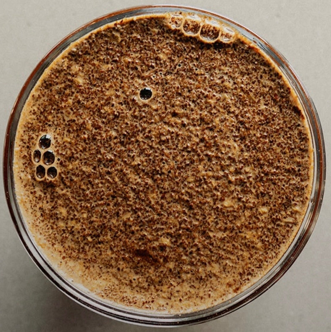 Top-down view of a frothy brown beverage in a clear glass, showing a thick layer of foam on a neutral gray background.