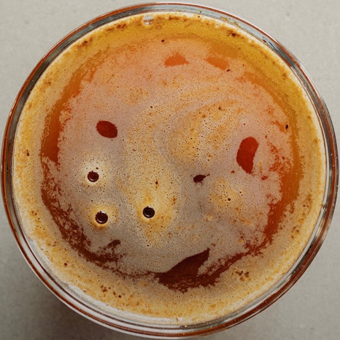 Top-down view of a clear glass filled with an orange-brown beverage, showing a foamy surface with small dark bubbles, set against a plain light gray background.