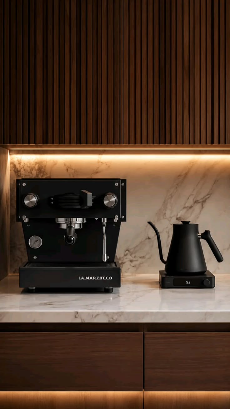 A black La Marzocco espresso machine sits on a white marble countertop, with a matching matte black gooseneck kettle on a small base to the right. The background features a warm wood slat wall and a marble backsplash illuminated by warm under-cabinet lighting.