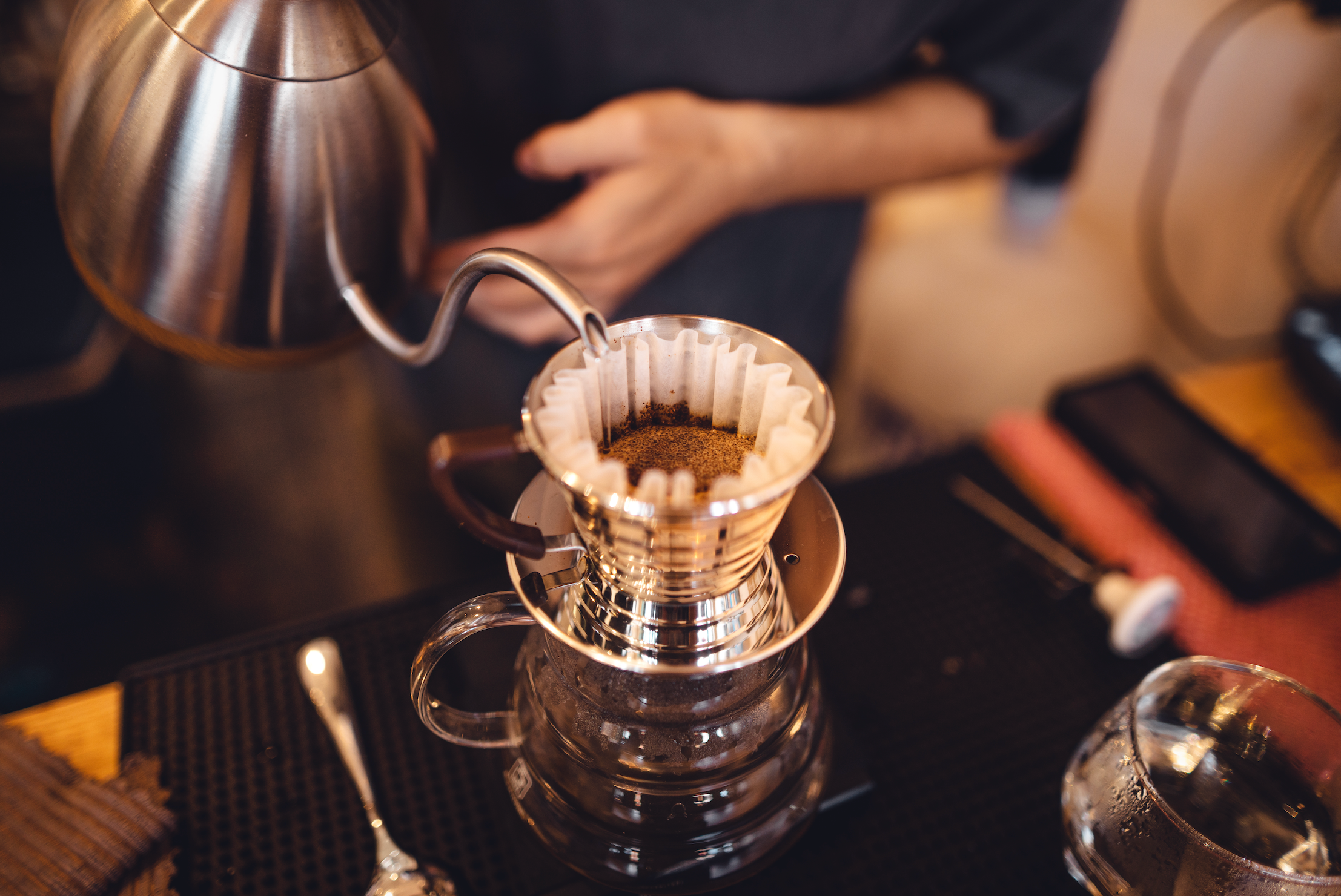 Person making coffee using a pour-over method with a metal kettle and glass carafe.